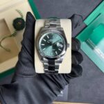 Rolex Datejust watch on a cream display cushion; green dial, date at 3, held by a gloved hand. Close-up of stainless Rolex Datejust with emerald-green dial and date window, on a beige cushion. Rolex Oyster Perpetual Datejust in stainless steel with green dial, shown on a display cushion with packaging in background. Watch detail: Rolex Datejust stainless bracelet, green dial, date magnifier, presented on a display stand.