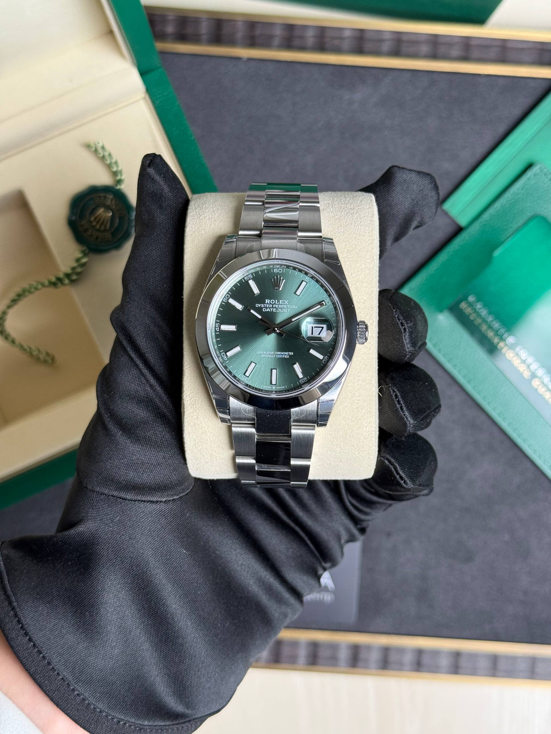 Rolex Datejust watch on a cream display cushion; green dial, date at 3, held by a gloved hand. Close-up of stainless Rolex Datejust with emerald-green dial and date window, on a beige cushion. Rolex Oyster Perpetual Datejust in stainless steel with green dial, shown on a display cushion with packaging in background. Watch detail: Rolex Datejust stainless bracelet, green dial, date magnifier, presented on a display stand.