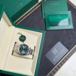 Rolex watch displayed in an open beige inner box with a green outer box, alongside a green warranty card and booklet.