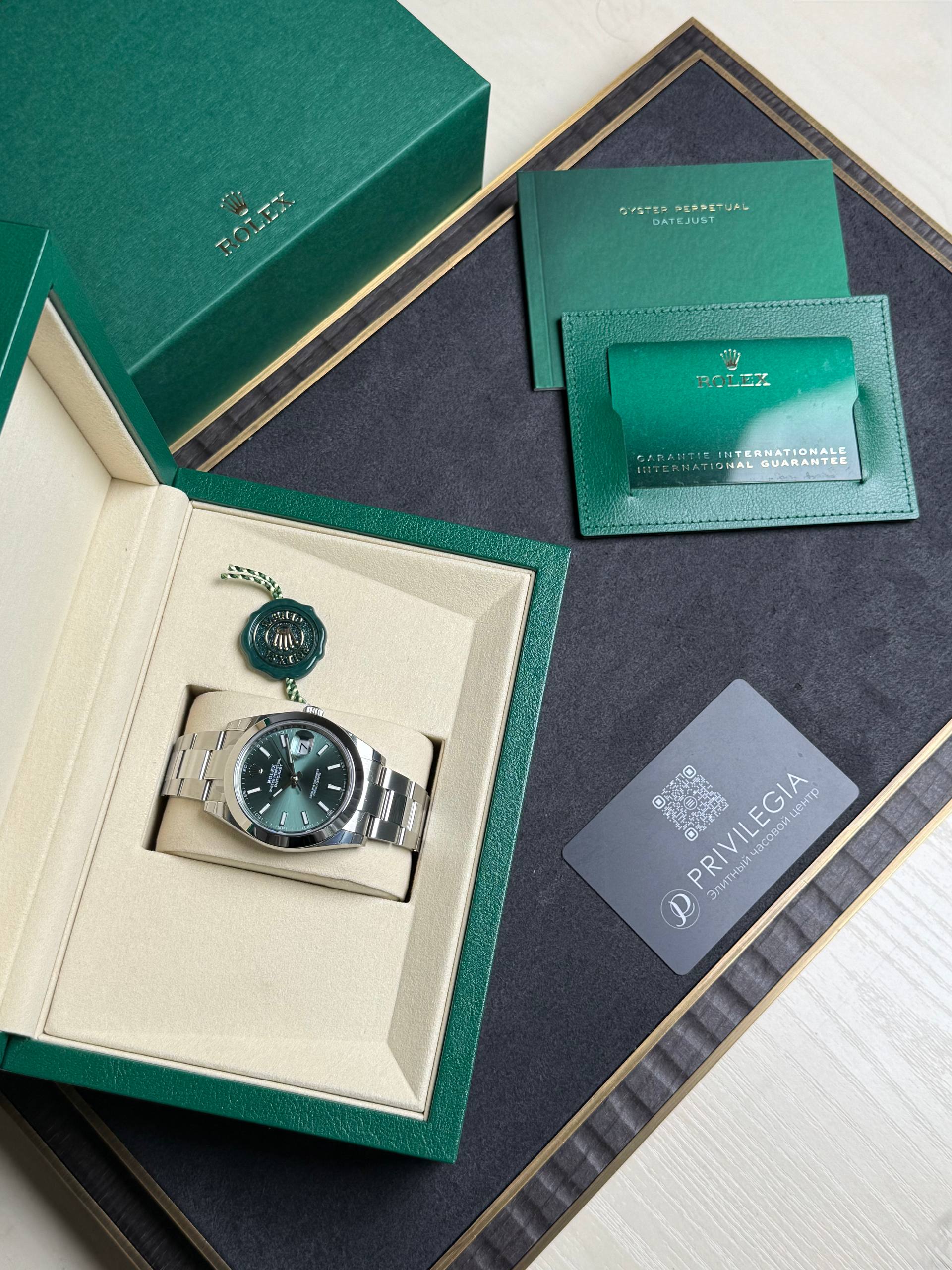 Rolex watch displayed in an open beige inner box with a green outer box, alongside a green warranty card and booklet.