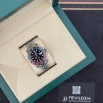 Luxury stainless steel watch with black dial and red/blue bezel in a green presentation box.