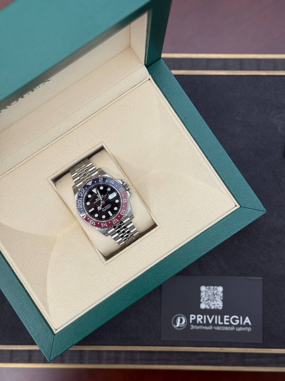 Luxury stainless steel watch with black dial and red/blue bezel in a green presentation box.