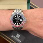 Rolex GMT-Master II with a red/blue bezel on a metal Jubilee bracelet worn on the left wrist; black dial with date magnifier.