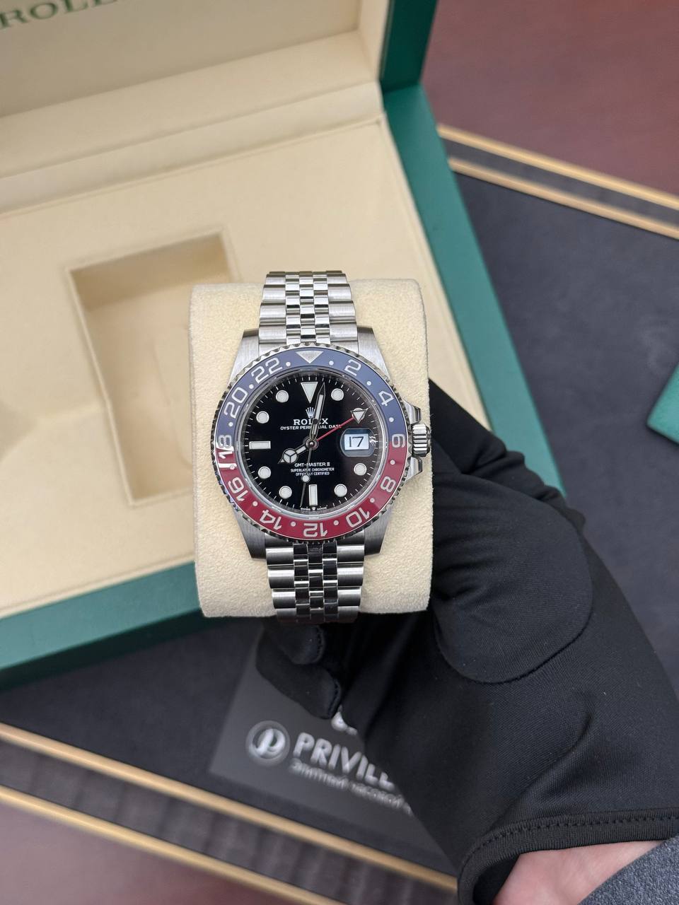 Rolex GMT-Master II with black dial, red/blue bezel, and Jubilee bracelet held by a gloved hand in a beige presentation box.