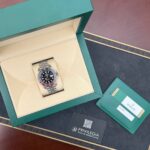 Rolex watch with stainless bracelet in a green presentation box, beige interior, and accompanying green warranty card on a black surface.