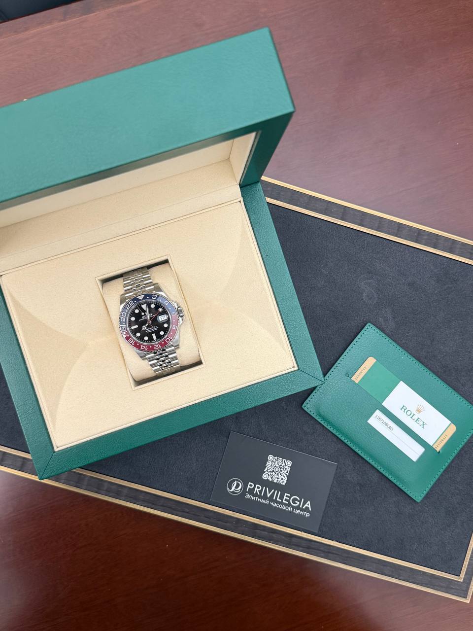 Rolex watch with stainless bracelet in a green presentation box, beige interior, and accompanying green warranty card on a black surface.
