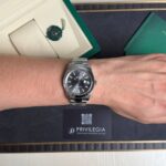 Silver Rolex watch on a forearm, with green Rolex box, green booklet, and a Privilegia card visible in the background.