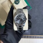 Silver Rolex Oyster Perpetual Datejust wristwatch on a beige display cushion, held by a gloved hand; box and green edge in the background.