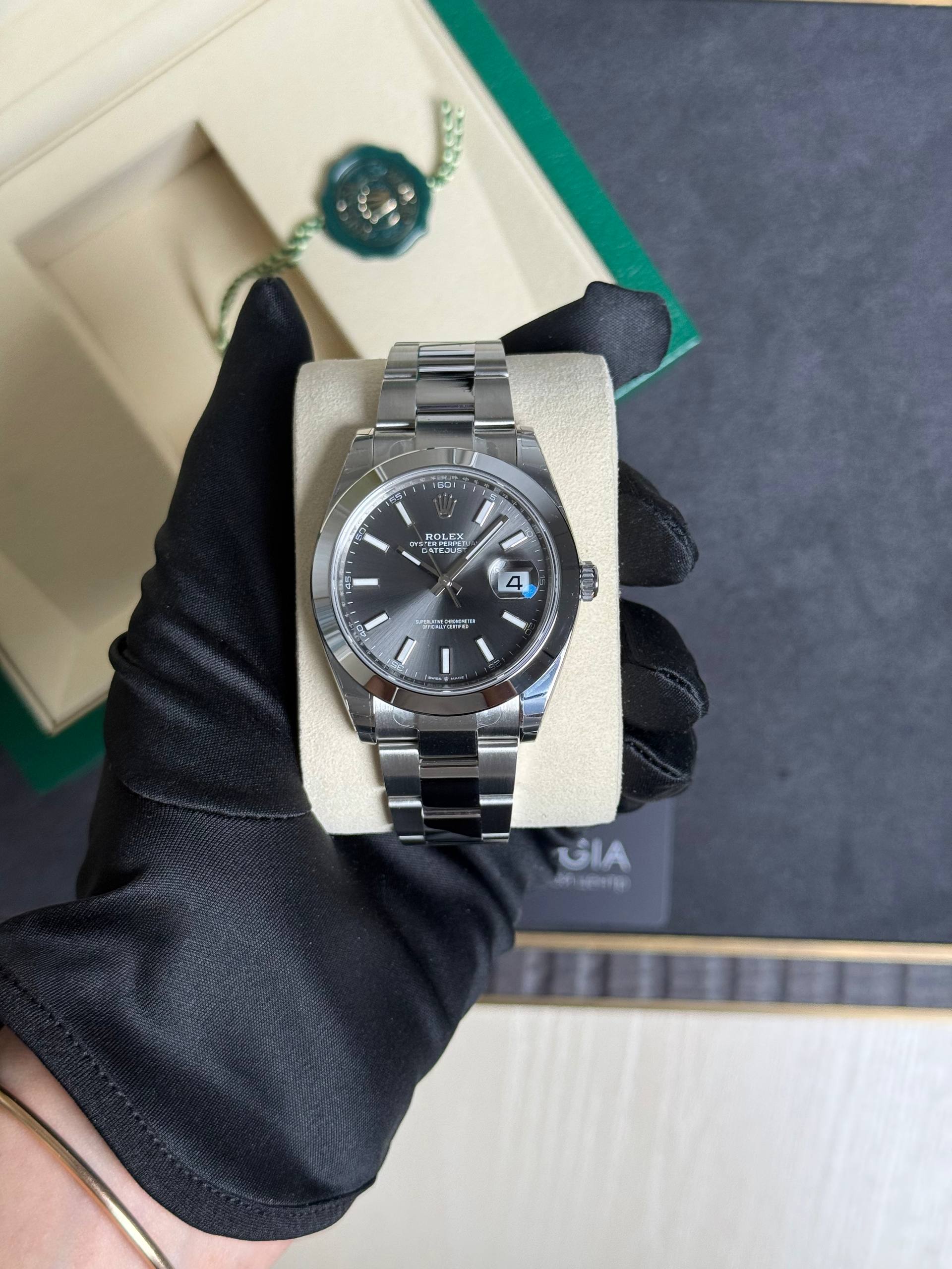 Silver Rolex Oyster Perpetual Datejust wristwatch on a beige display cushion, held by a gloved hand; box and green edge in the background.