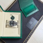 Rolex watch displayed in a beige insert inside a green presentation box, with a green warranty card and booklet nearby.