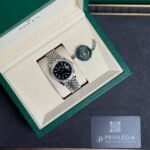 Rolex watch in an open green box with beige interior and a green seal tag beside it.