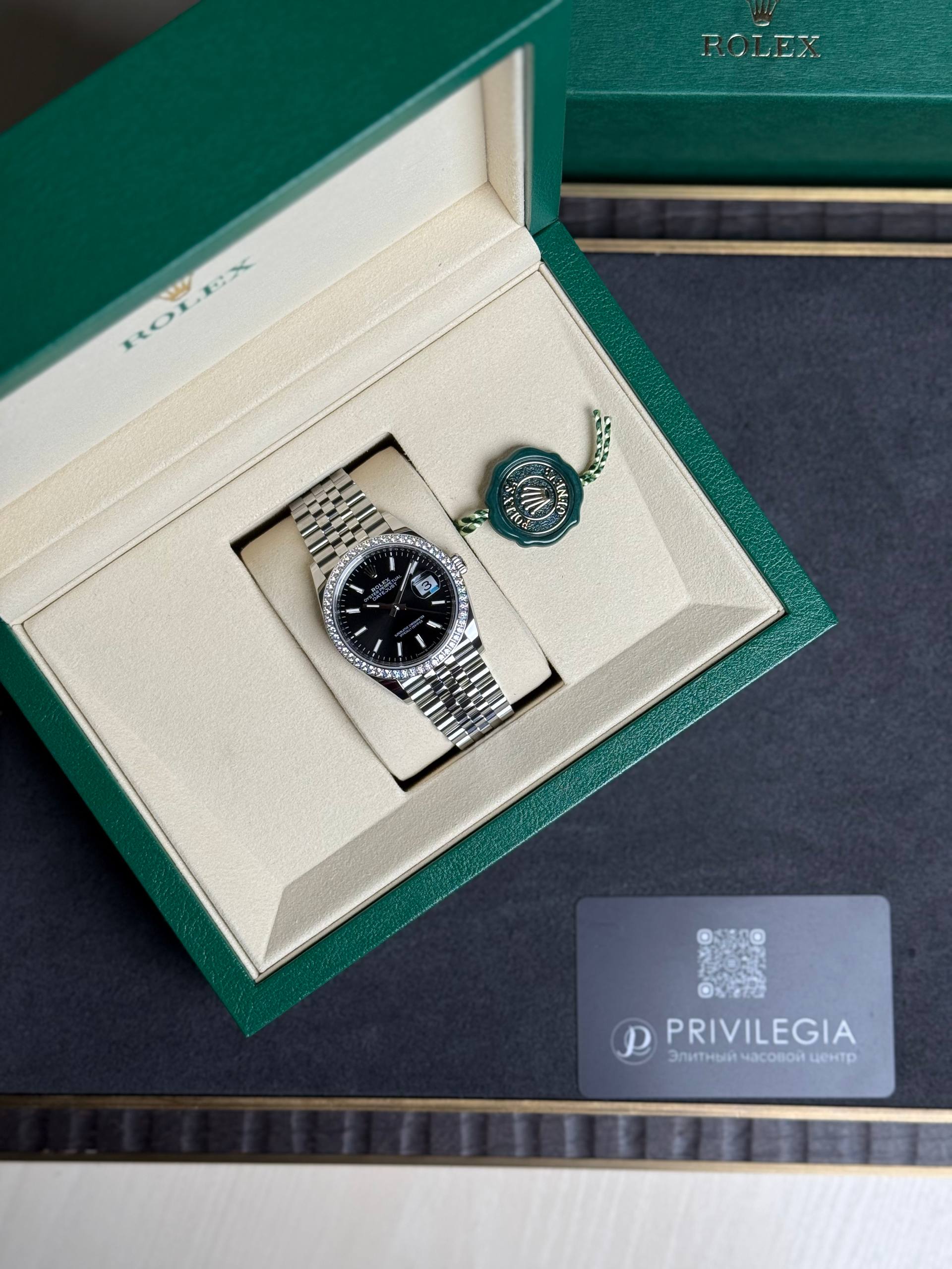Rolex watch in an open green box with beige interior and a green seal tag beside it.
