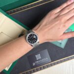Silver Rolex watch with black face and diamond bezel on the wrist, inside an open green Rolex box.