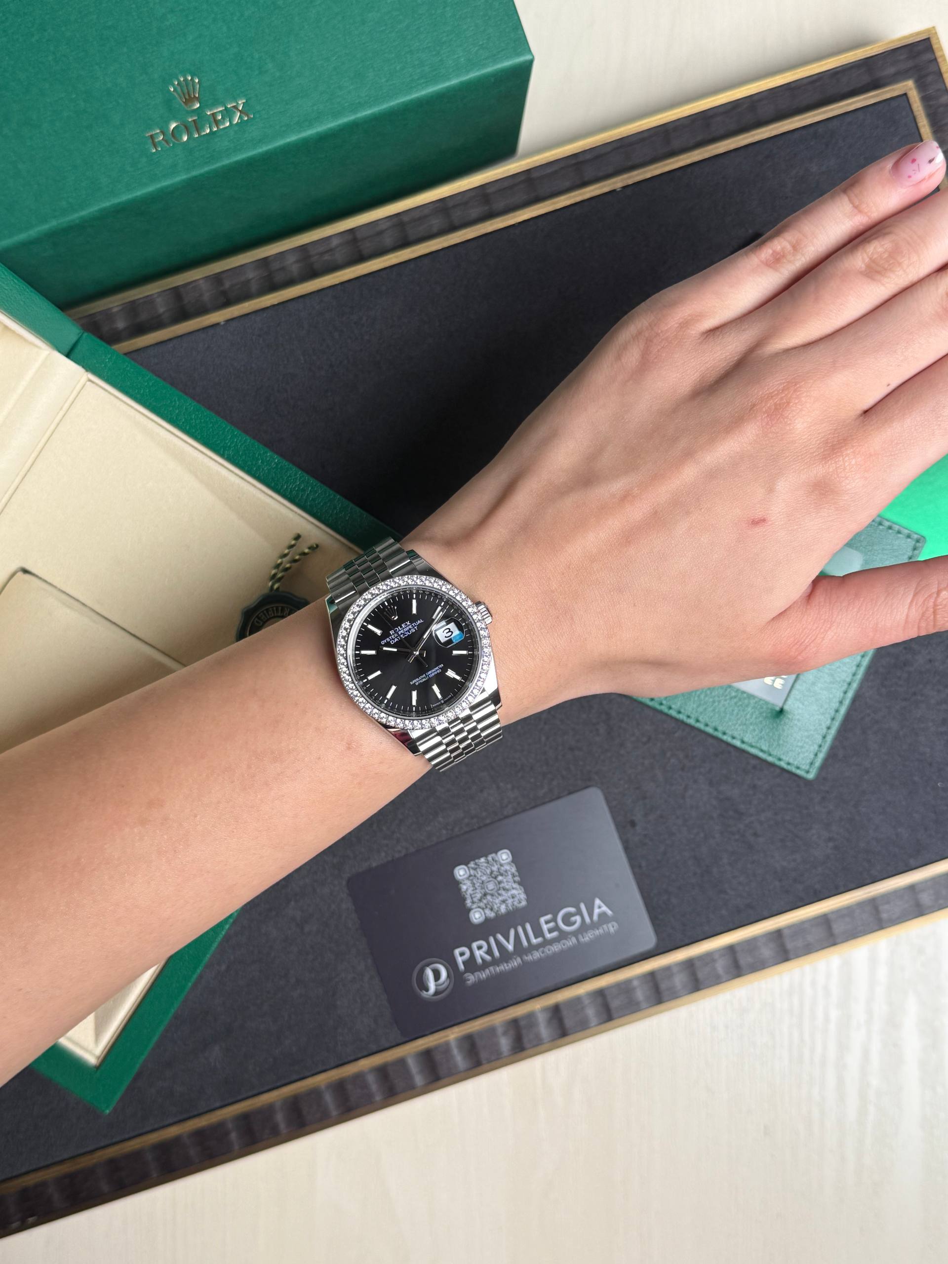 Silver Rolex watch with black face and diamond bezel on the wrist, inside an open green Rolex box.