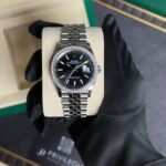 Rolex Datejust with diamond bezel and black dial, date window at 3, in a beige display box held by a gloved hand.