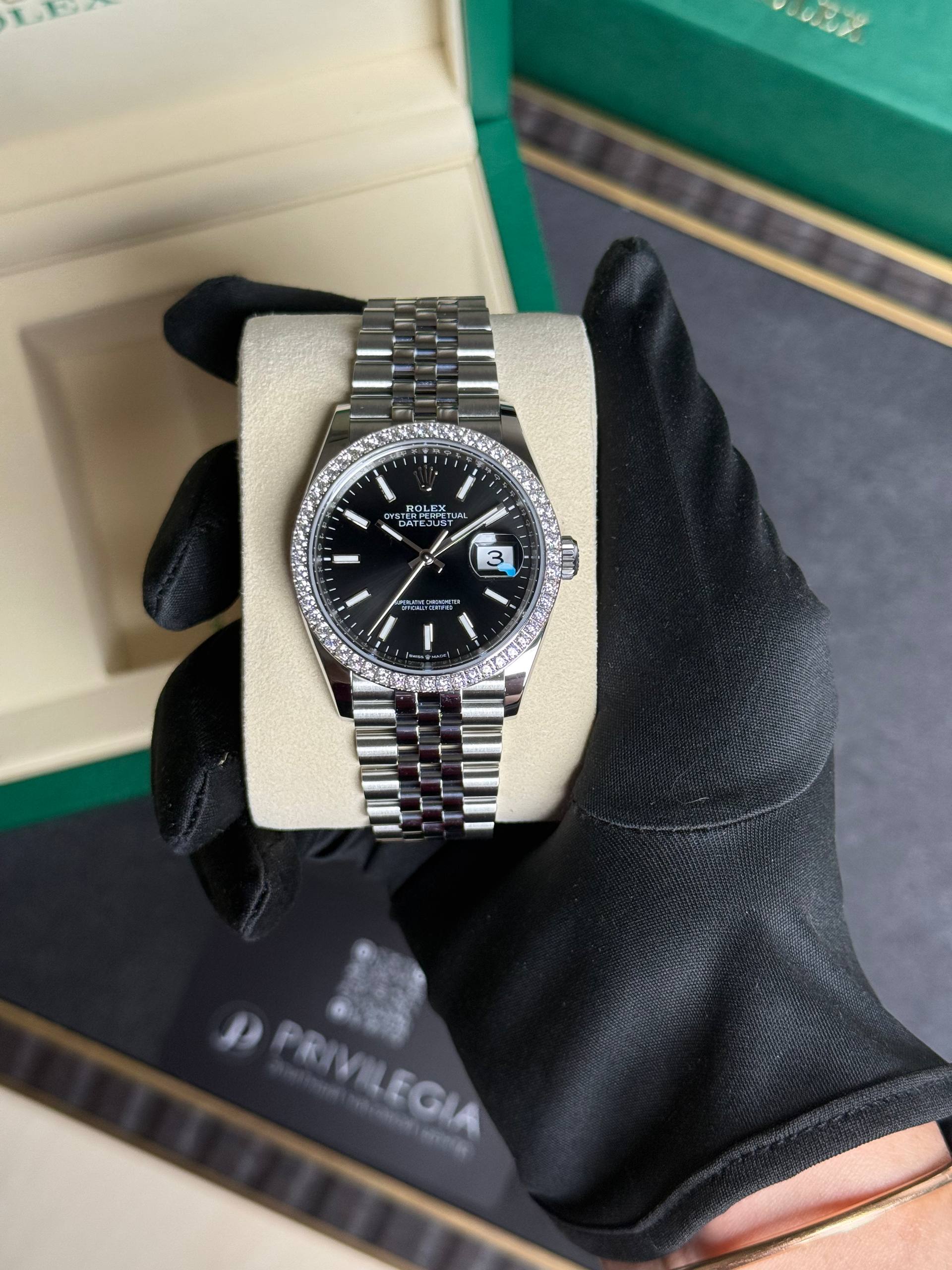 Rolex Datejust with diamond bezel and black dial, date window at 3, in a beige display box held by a gloved hand.