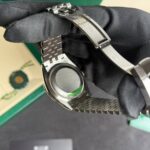 Close-up of a stainless steel watch band with a deployment clasp, held by a gloved hand, green box in the background.