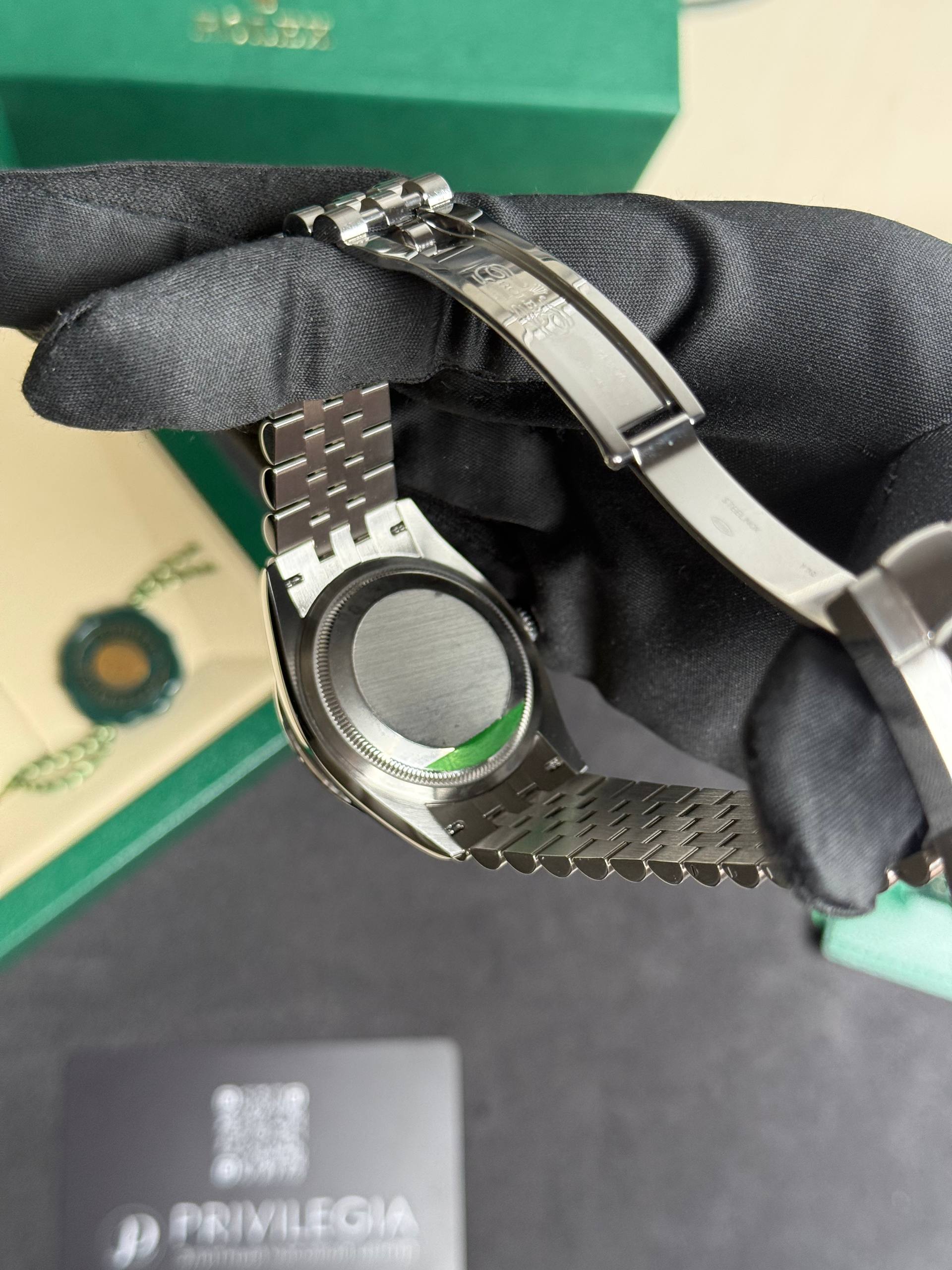 Close-up of a stainless steel watch band with a deployment clasp, held by a gloved hand, green box in the background.