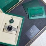 Rolex watch in a beige display box inside a green presentation box, with a green warranty card and documents nearby.