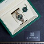 Rolex watch with gold-and-silver bracelet in a green presentation box, alongside a green seal tag.