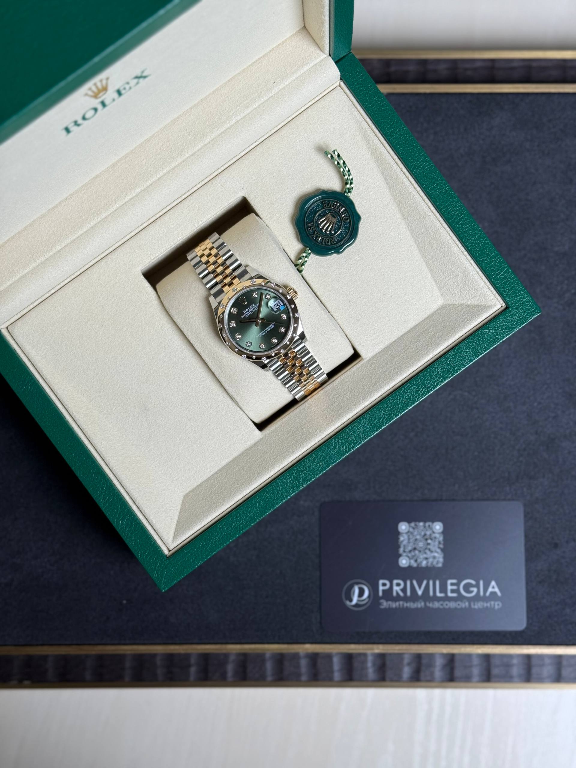 Rolex watch with gold-and-silver bracelet in a green presentation box, alongside a green seal tag.