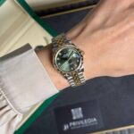 Rolex Datejust wristwatch with a green dial, diamond hour markers, and a two-tone gold and steel bracelet on a wrist beside a green-edged box.