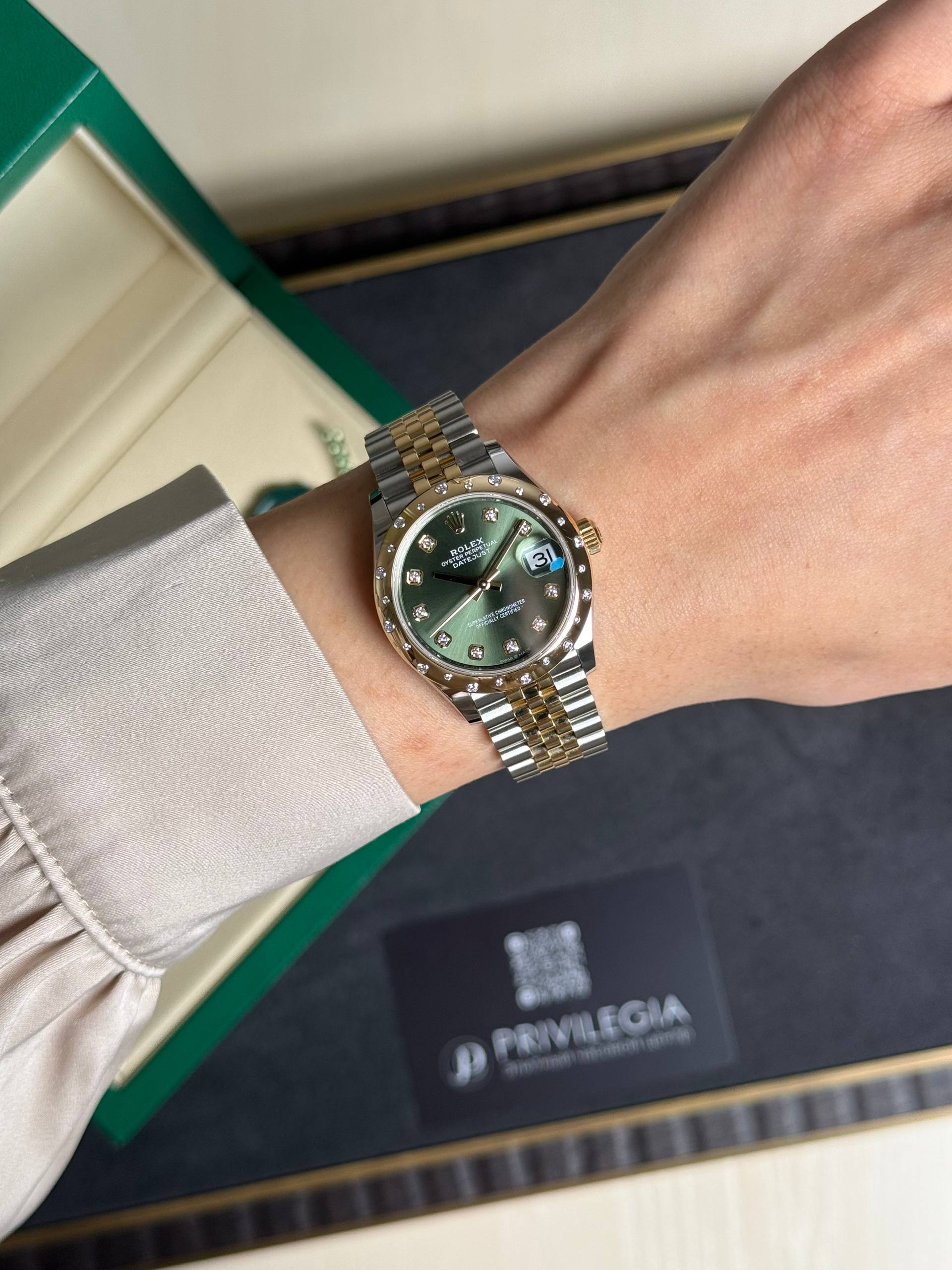 Rolex Datejust wristwatch with a green dial, diamond hour markers, and a two-tone gold and steel bracelet on a wrist beside a green-edged box.