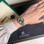 Wrist wearing a two-tone Rolex watch on a beige-sleeved arm resting on an open green presentation box with a black interior and card nearby