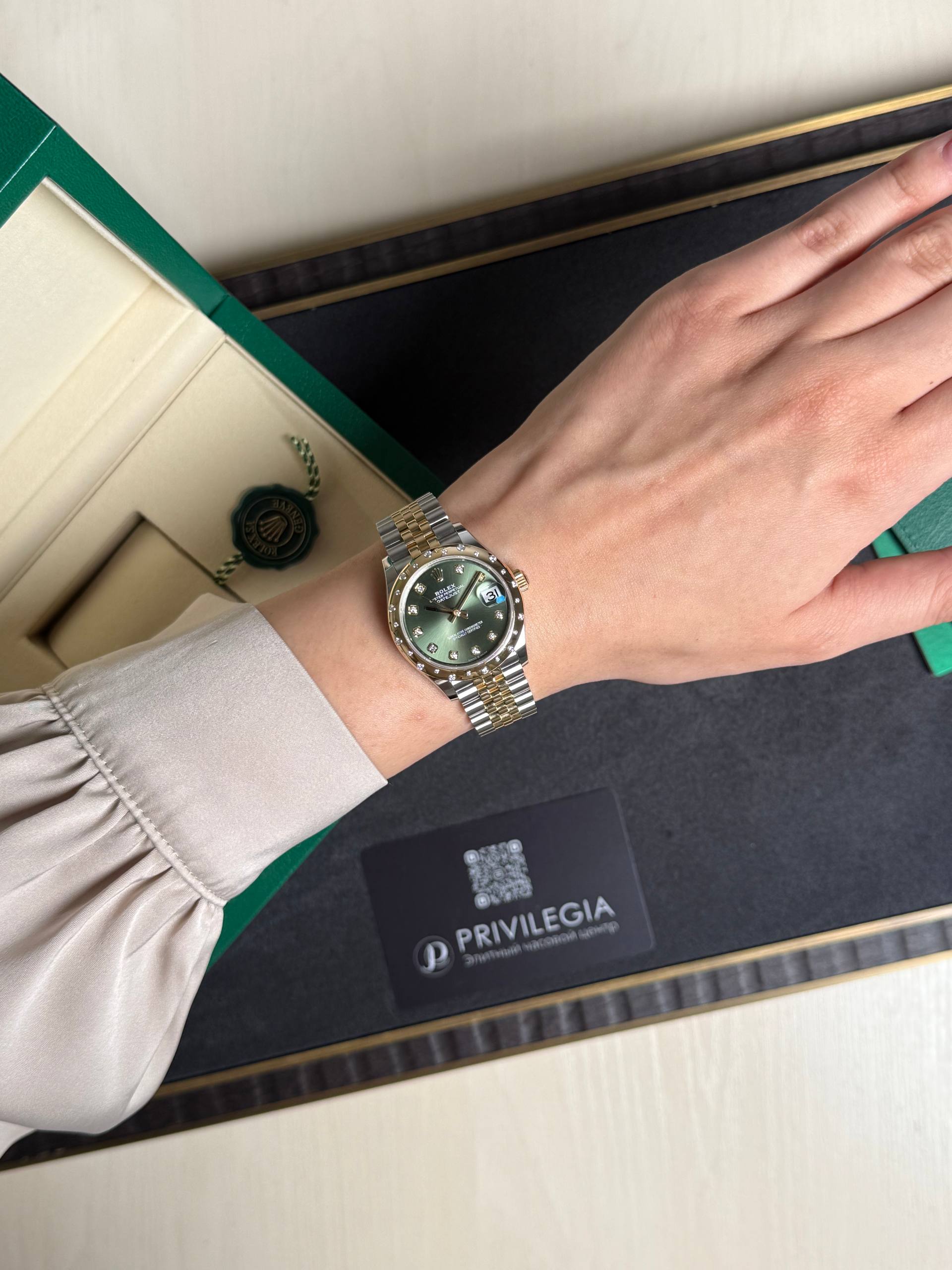 Wrist wearing a two-tone Rolex watch on a beige-sleeved arm resting on an open green presentation box with a black interior and card nearby