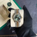 Two-tone Rolex Datejust with green dial and diamond hour markers on a padded display held by a gloved hand.