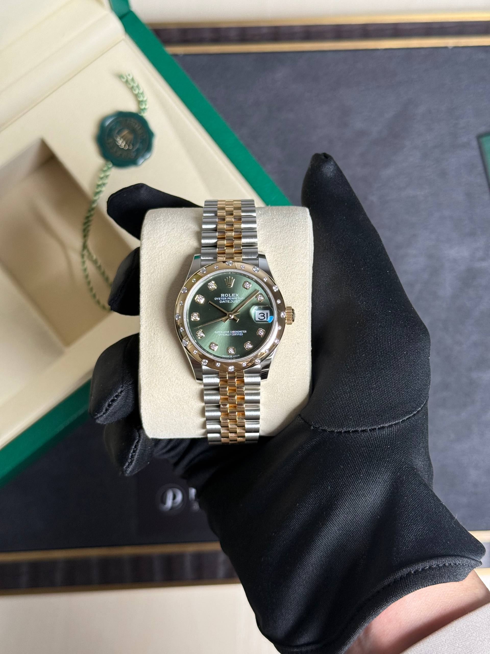 Two-tone Rolex Datejust with green dial and diamond hour markers on a padded display held by a gloved hand.