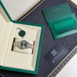 Rolex watch in beige presentation box with green outer case, along with green warranty cards on a dark surface.