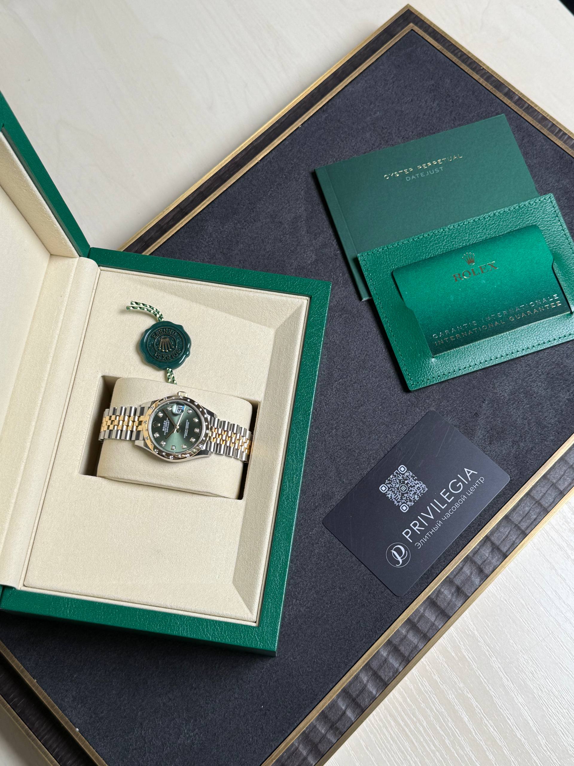 Rolex watch in beige presentation box with green outer case, along with green warranty cards on a dark surface.
