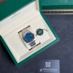 Open green Rolex box displaying a stainless steel Rolex watch with blue dial and matching seal tag beside it
