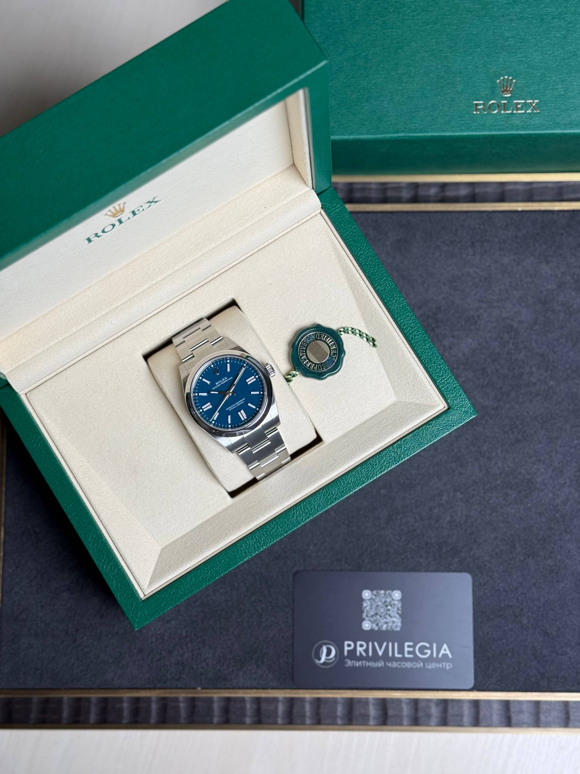 Open green Rolex box displaying a stainless steel Rolex watch with blue dial and matching seal tag beside it