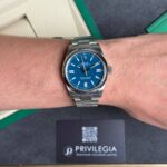 Wrist wearing a silver Rolex with a blue dial, metal bracelet, close-up shot; a black Privilegia card is visible in the background on a dark surface.