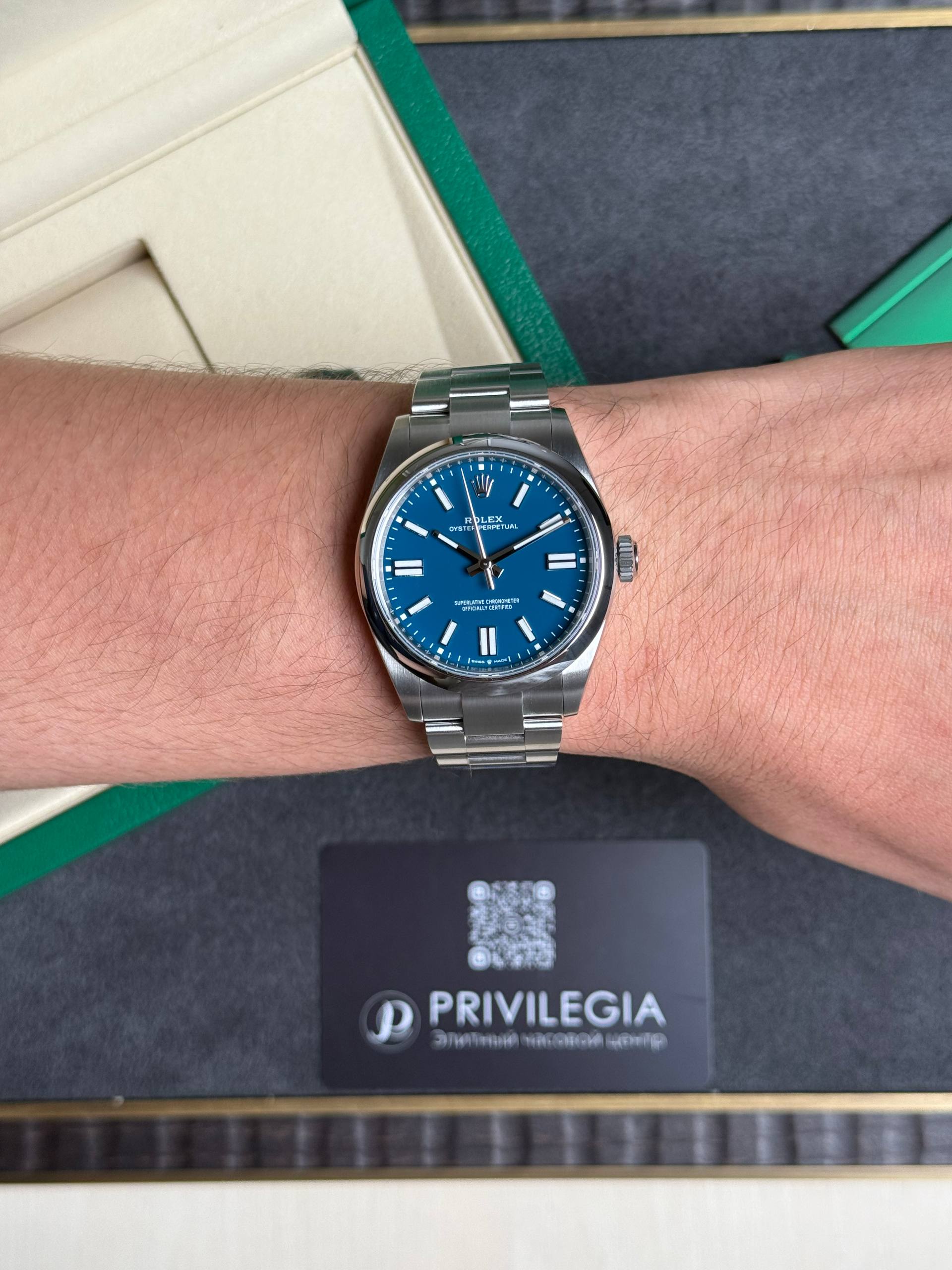 Wrist wearing a silver Rolex with a blue dial, metal bracelet, close-up shot; a black Privilegia card is visible in the background on a dark surface.