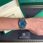 Wrist wearing a stainless steel Rolex with a blue dial, foregrounded by green Rolex boxes and a Privilegia card in the background