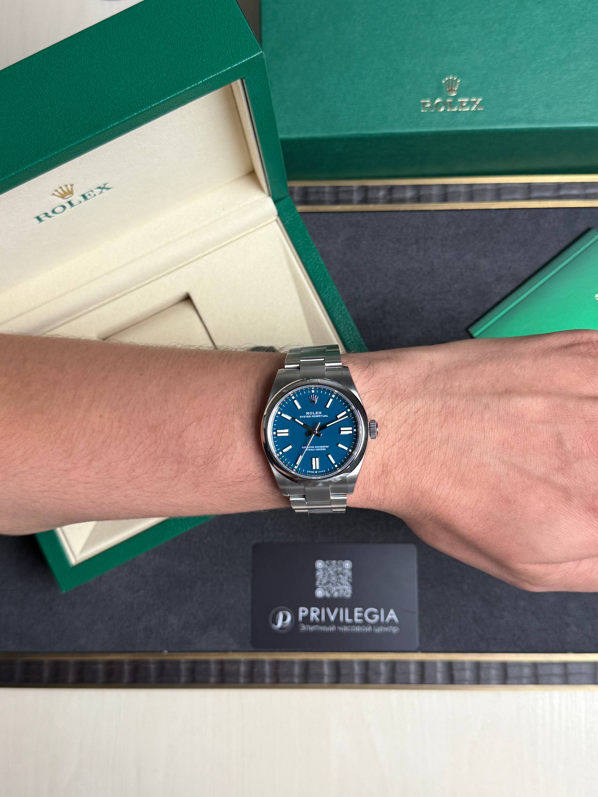 Wrist wearing a stainless steel Rolex with a blue dial, foregrounded by green Rolex boxes and a Privilegia card in the background