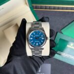 Rolex Oyster Perpetual on a beige cushion, blue dial, held by a gloved hand beside a green Rolex box in the background.