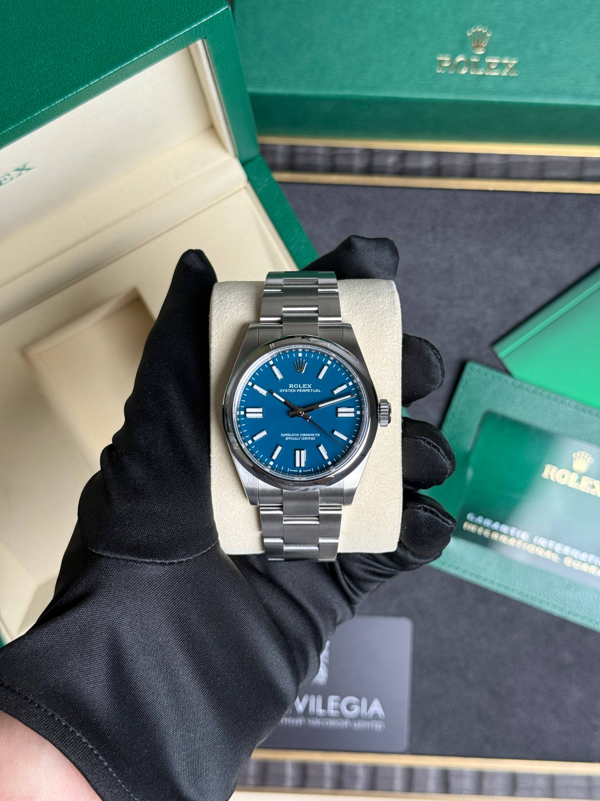 Rolex Oyster Perpetual on a beige cushion, blue dial, held by a gloved hand beside a green Rolex box in the background.