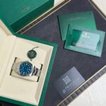 Rolex watch displayed in a beige insert inside a green box, with green warranty cards nearby much of the packaging visible.