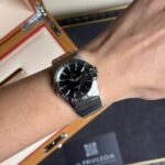 Wrist wearing an Omega Constellation stainless steel watch with black dial and date window, background shows a wooden watch box.