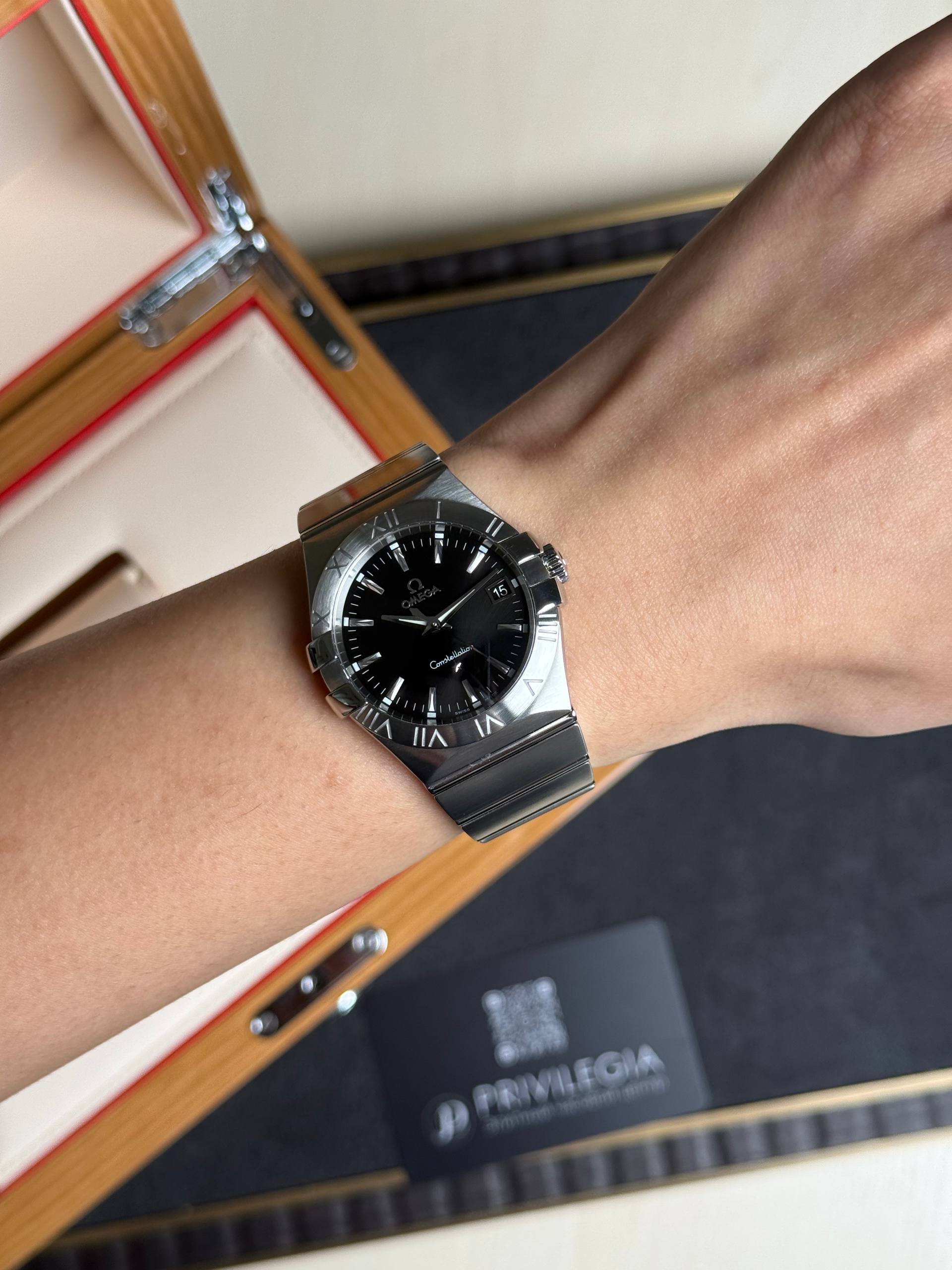 Wrist wearing an Omega Constellation stainless steel watch with black dial and date window, background shows a wooden watch box.