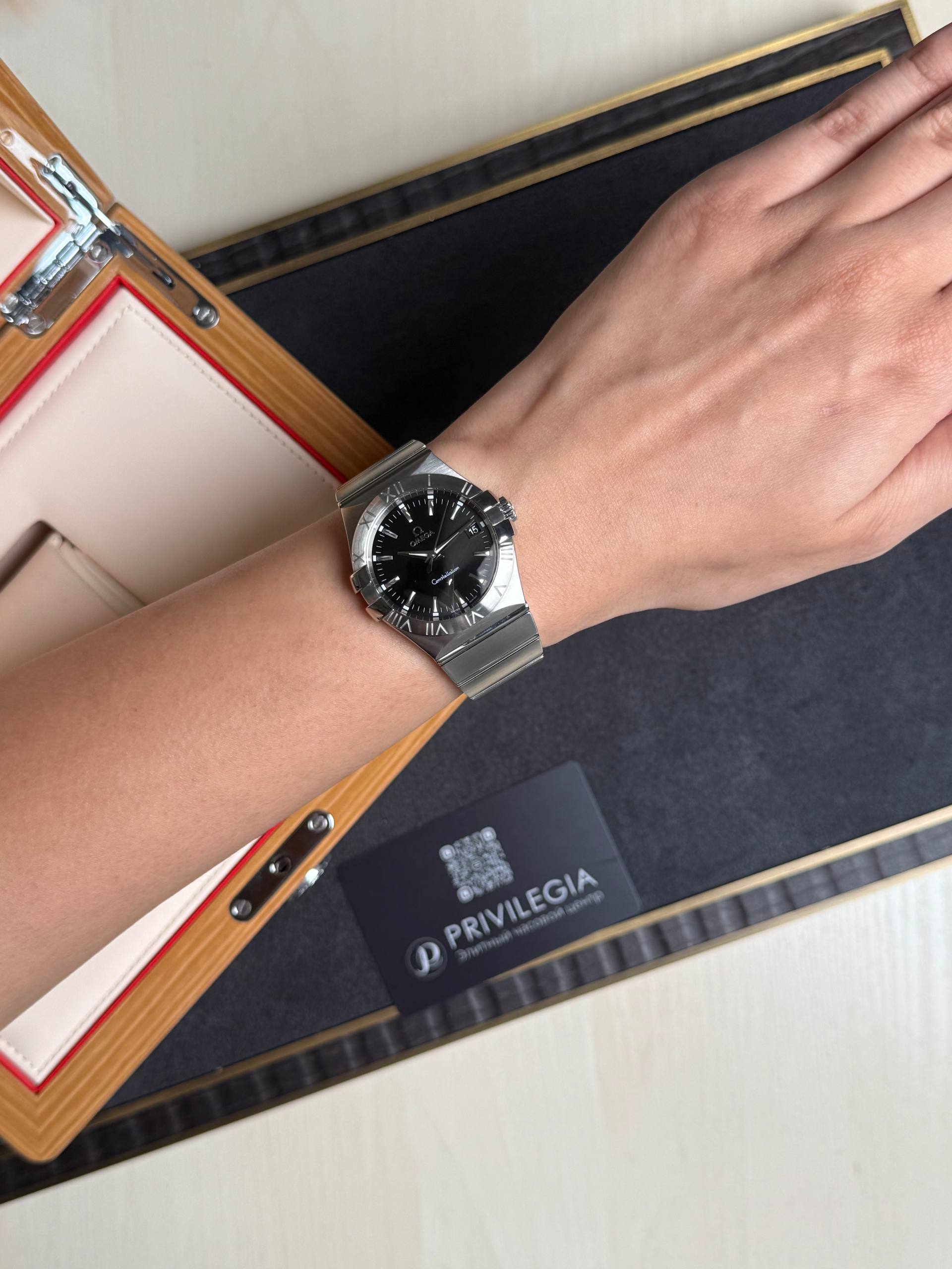 Close-up of a wrist wearing a silver metal watch with a black dial and Roman numerals, resting near a watch box with beige and red lining in the background.