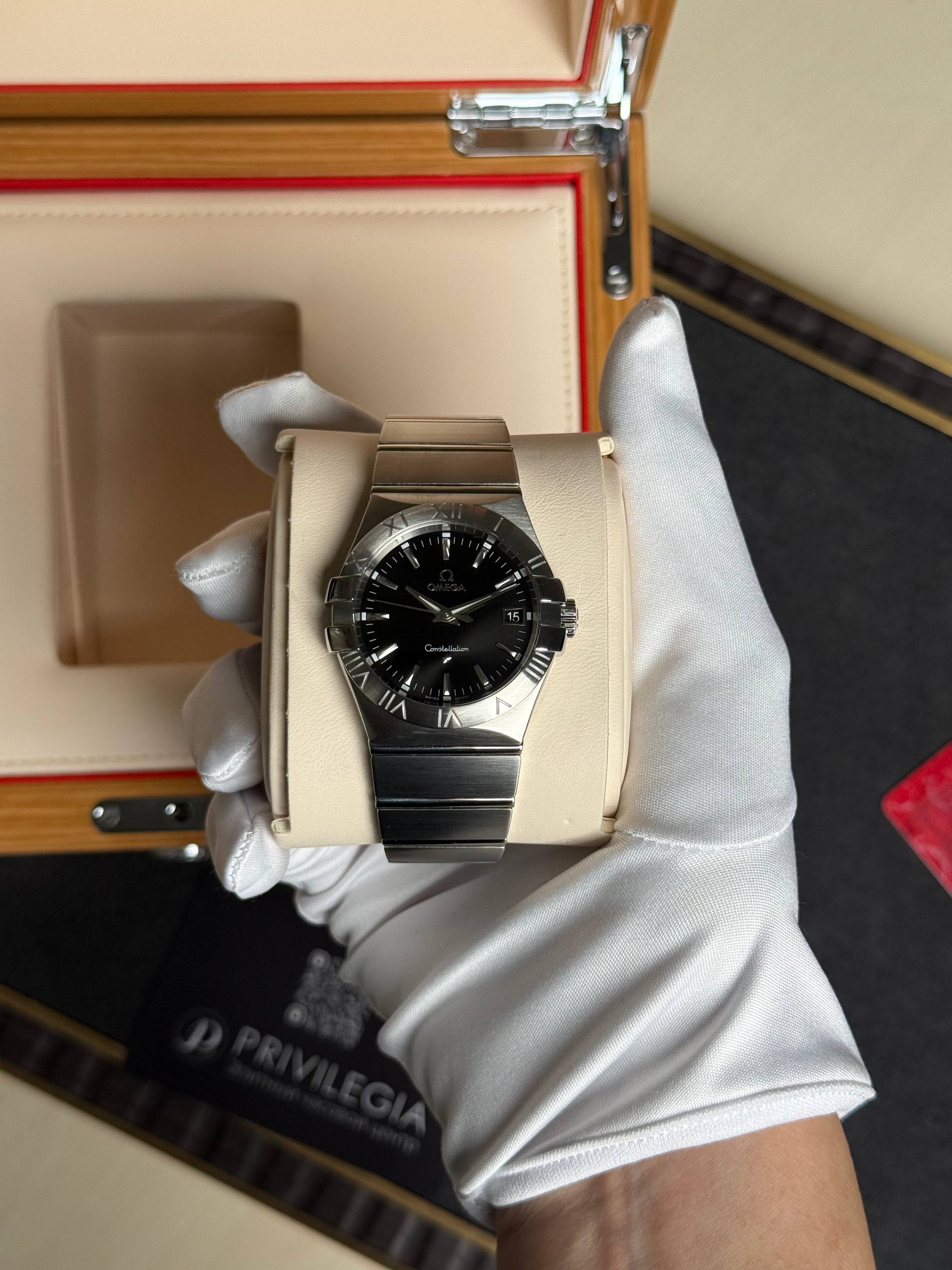 Omega Constellation wristwatch in a light beige display box with white gloves holding it, close-up view.