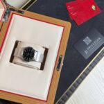 Wristwatch in an open beige cushioned display case with a wooden frame, beside red warranty cards on a dark surface.