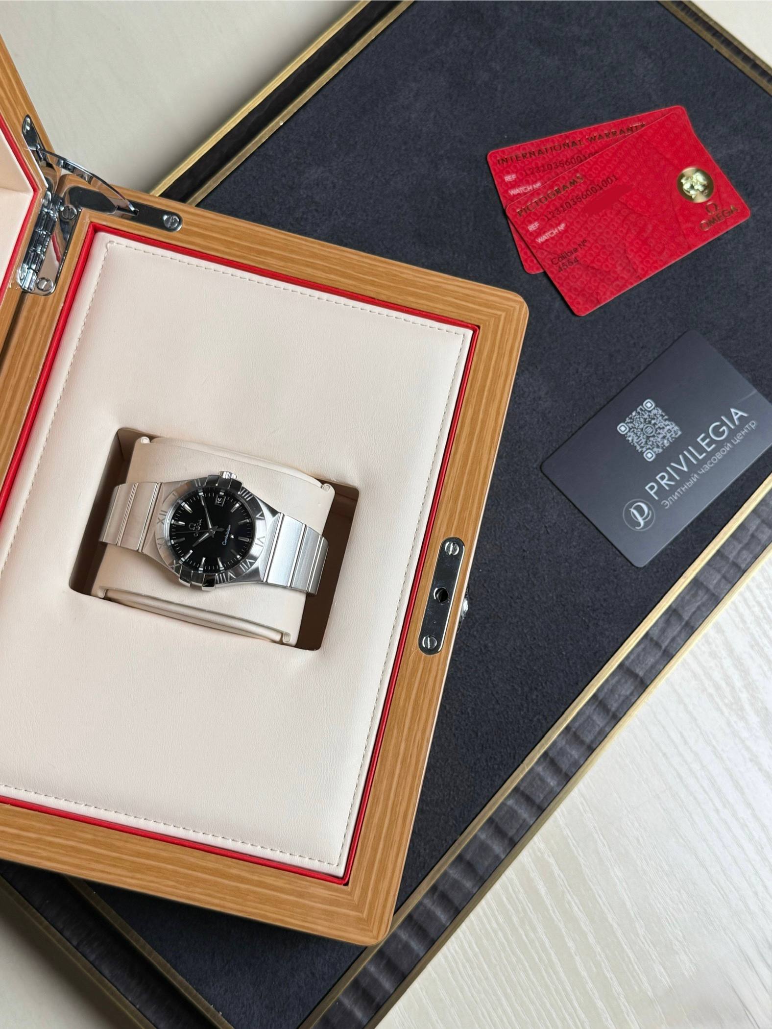 Wristwatch in an open beige cushioned display case with a wooden frame, beside red warranty cards on a dark surface.