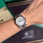 Silver Rolex Datejust wristwatch with diamond hour markers on a metal bracelet worn on the wrist, green Rolex boxes in the background
