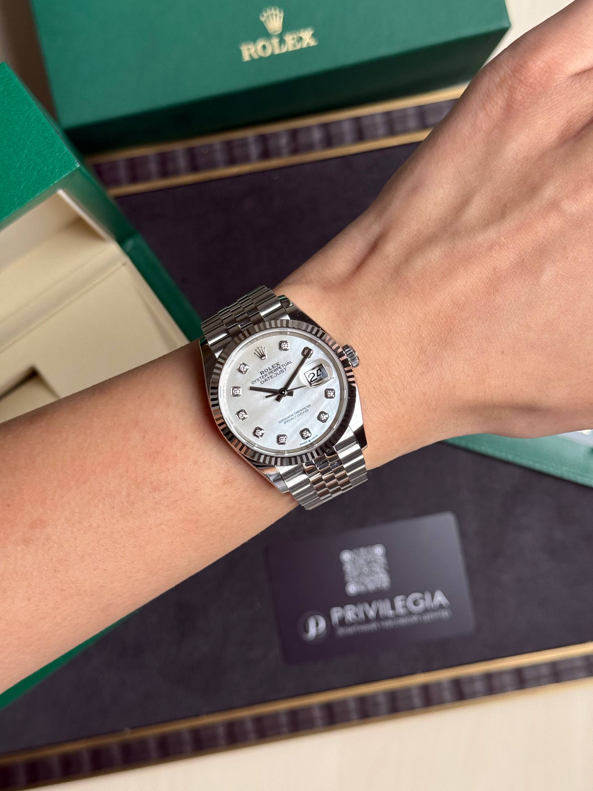 Silver Rolex Datejust wristwatch with diamond hour markers on a metal bracelet worn on the wrist, green Rolex boxes in the background